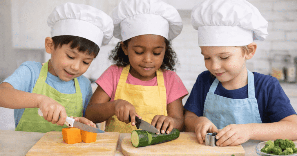 Three young children in chef hats and aprons cutting vegetables into geometric shapes like cubes, circles, and stars during a STEM in the Kitchen activity.