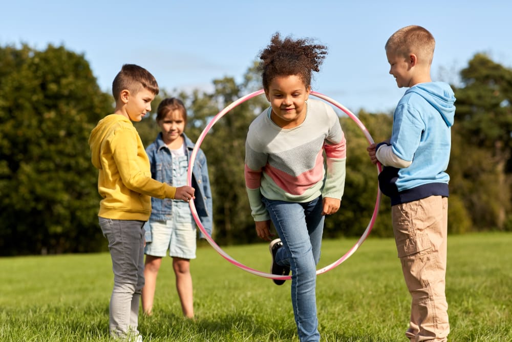 Diverse group of children playing a hula hoop team-building game outdoors to promote Social Emotional Learning (SEL) and physical wellness.
