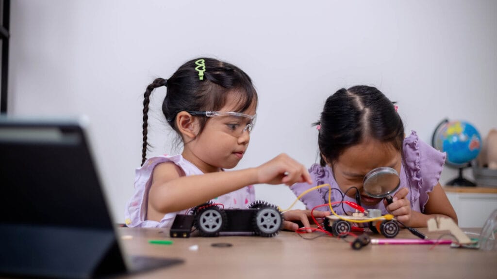Two young girls using a magnifying glass and tools to investigate robotic circuits and motors, building confidence through hands-on STEM mastering.