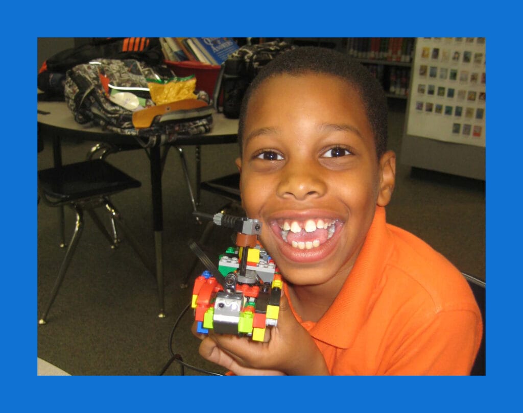 A smiling young boy in an orange polo shirt proudly holding a colorful LEGO robotics creation during an IntelliBricks after-school program.
