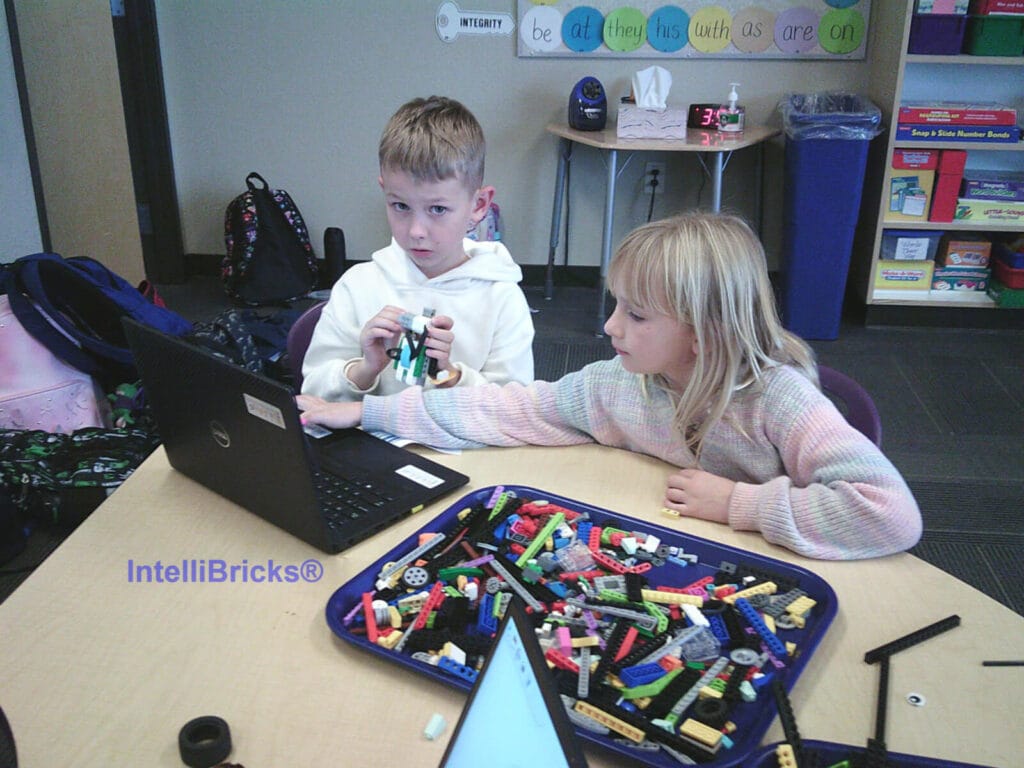 Two elementary students collaborating on a LEGO robotics build with a laptop and organized part tray.