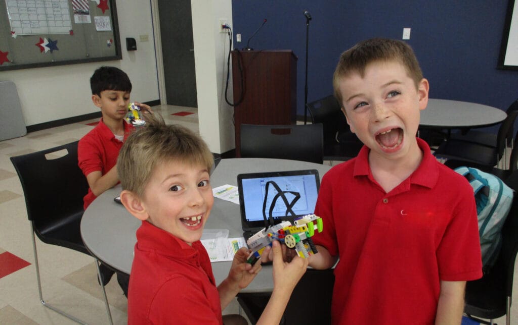 Two excited students in red shirts showing off their completed LEGO robotics project at summer camp.